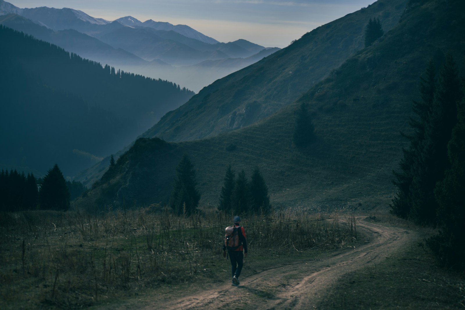 Lone hiker walking a mountain path toward distant peaks, where a hood and thumb loops on hiking hoodies keep the sun off